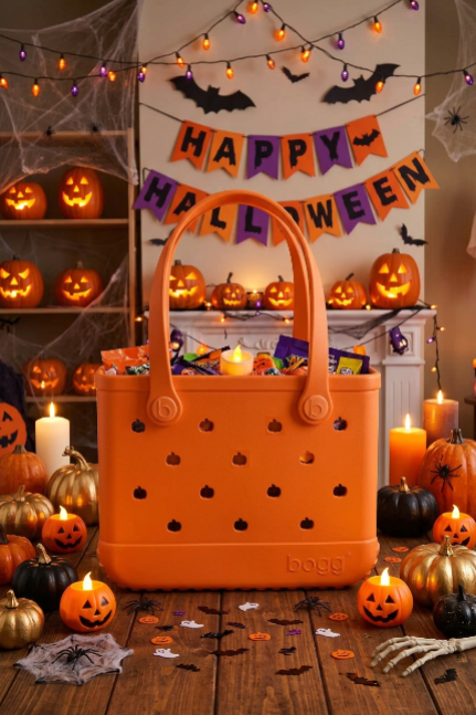 An orange Bogg Halloween basket filled with candy, set on a wooden floor in a decorated room with jack-o'-lanterns, candles, spiderwebs, and a 'Happy Halloween' banner.